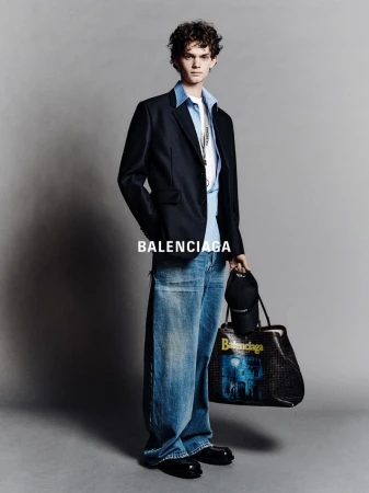 Full-length portrait of a young adult male wearing a black blazer, light blue button-down shirt, baggy jeans, and carrying a tote bag.