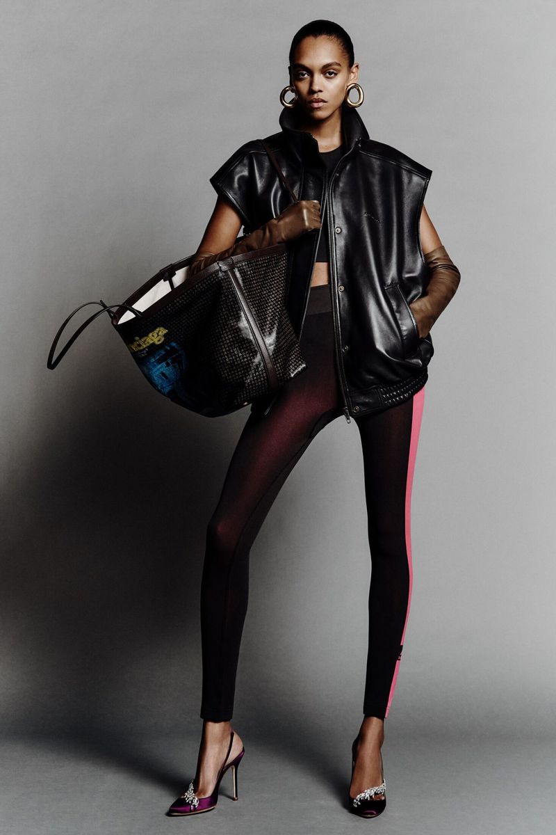 A stylish adult woman poses indoors wearing a black sleeveless vest, leggings with a pink stripe, heels, and gold hoop earrings.