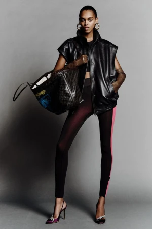 A stylish adult woman poses indoors wearing a black sleeveless vest, leggings with a pink stripe, heels, and gold hoop earrings.