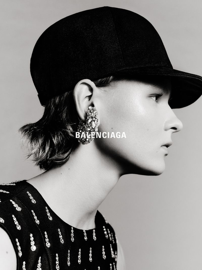 Close-up black-and-white portrait of an adult female wearing a black cap and ornate earrings with BALENCIAGA text overlayed on the image.