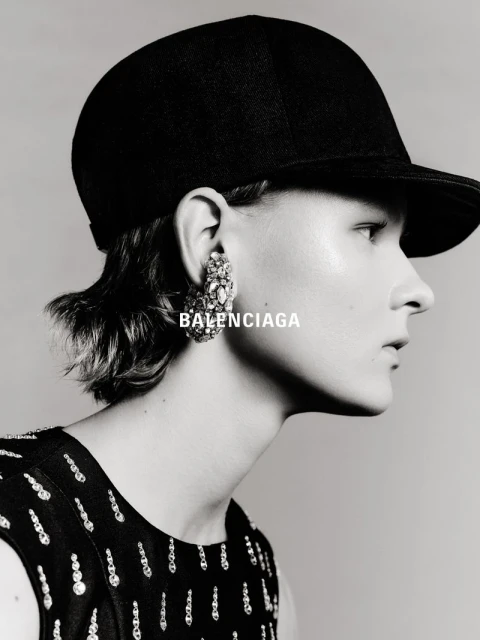 Close-up black-and-white portrait of an adult female wearing a black cap and ornate earrings with BALENCIAGA text overlayed on the image.