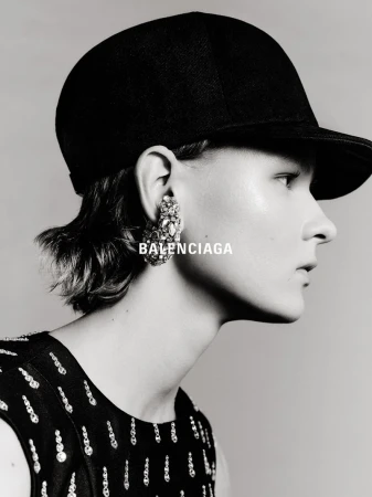 Close-up black-and-white portrait of an adult female wearing a black cap and ornate earrings with BALENCIAGA text overlayed on the image.