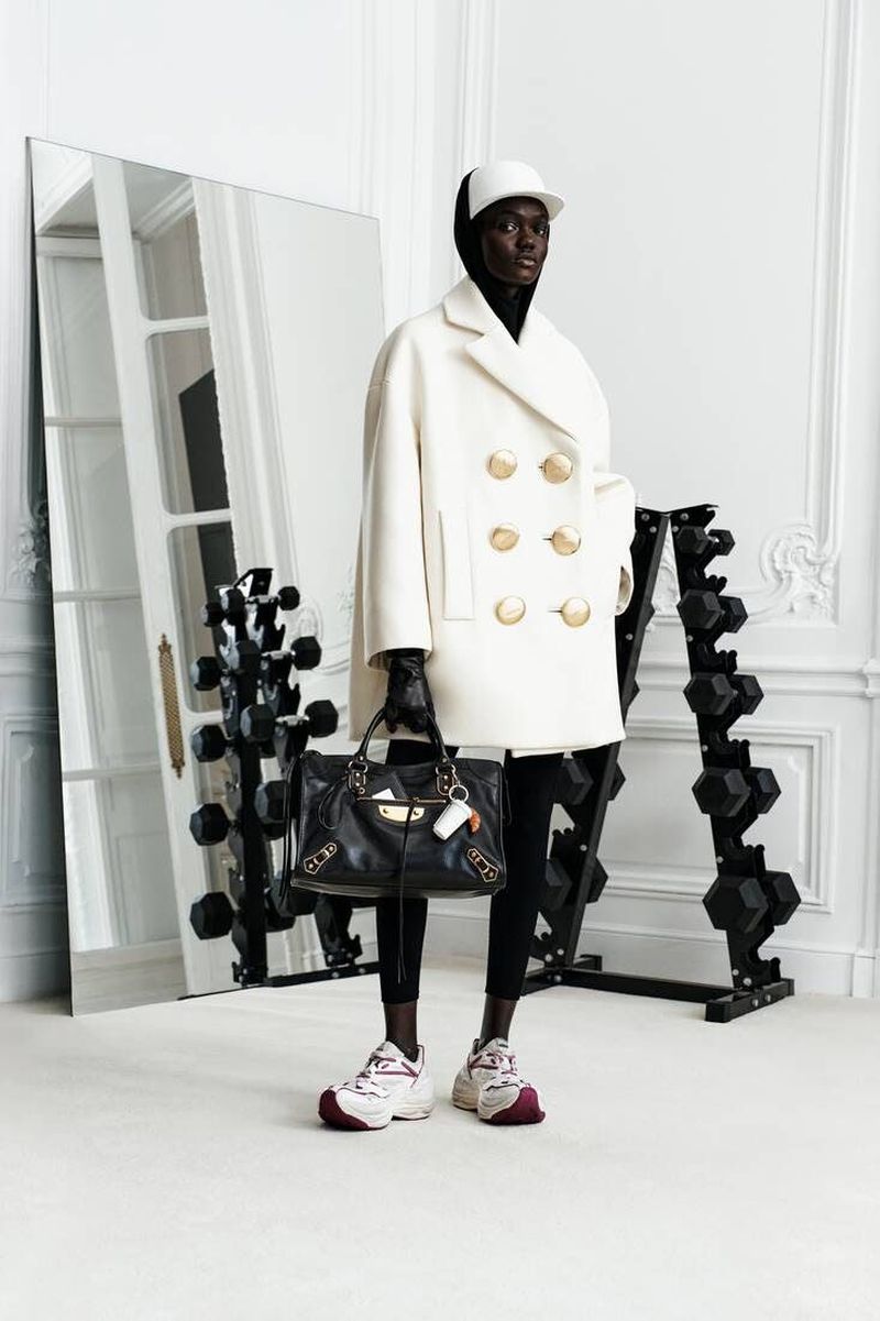 An adult fashion model poses indoors near weights and a mirror, wearing a white coat, white baseball cap, and black Balenciaga handbag.