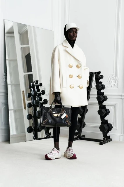 An adult fashion model poses indoors near weights and a mirror, wearing a white coat, white baseball cap, and black Balenciaga handbag.