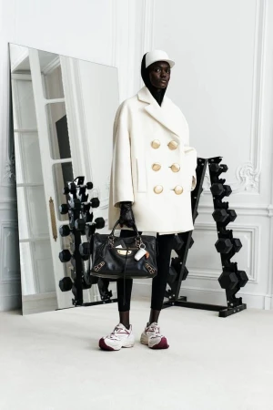 An adult fashion model poses indoors near weights and a mirror, wearing a white coat, white baseball cap, and black Balenciaga handbag.