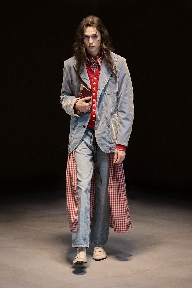 A person in a light blue jacket, red and white checkered shirt, and jeans, is holding a book while posing against a dark background.