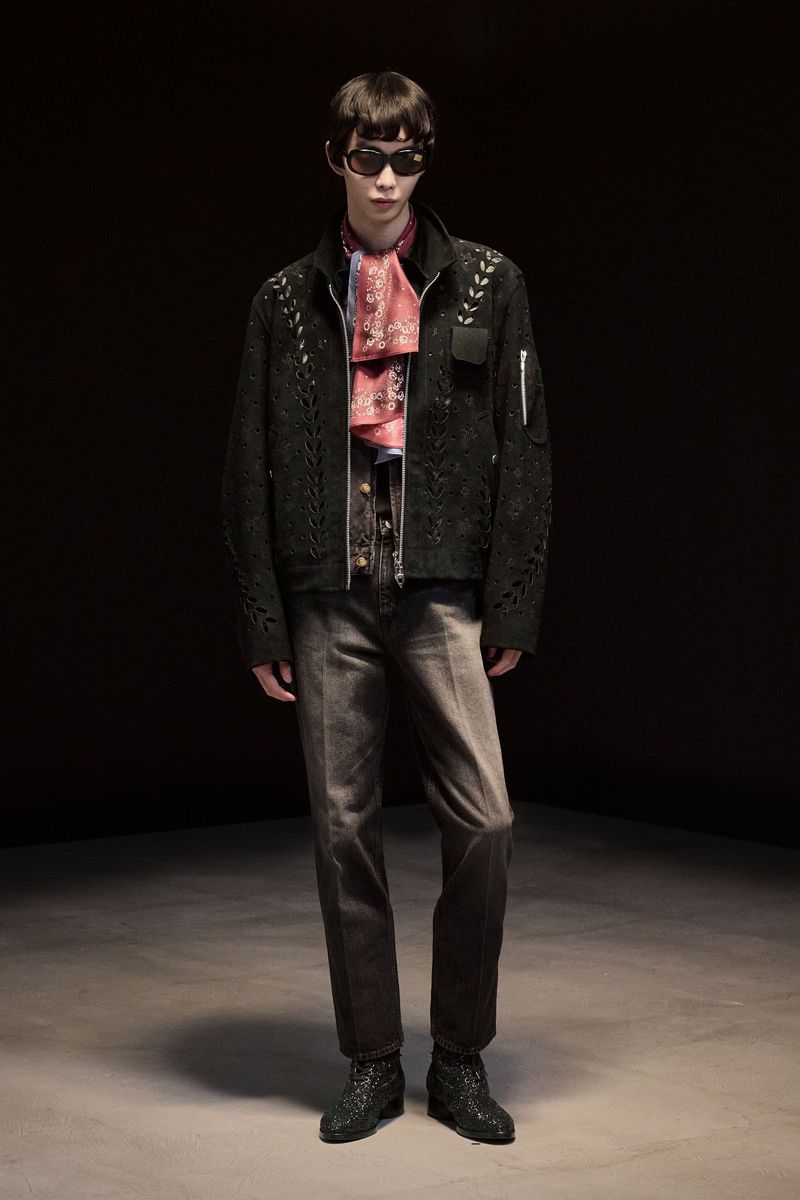 Male fashion model in a black jacket, grey jeans, sunglasses and patterned scarf poses in a dimly lit studio with a neutral floor.