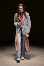 Model with long dark hair wearing a light blue blazer over a red cardigan and checkered fabric walking forward.