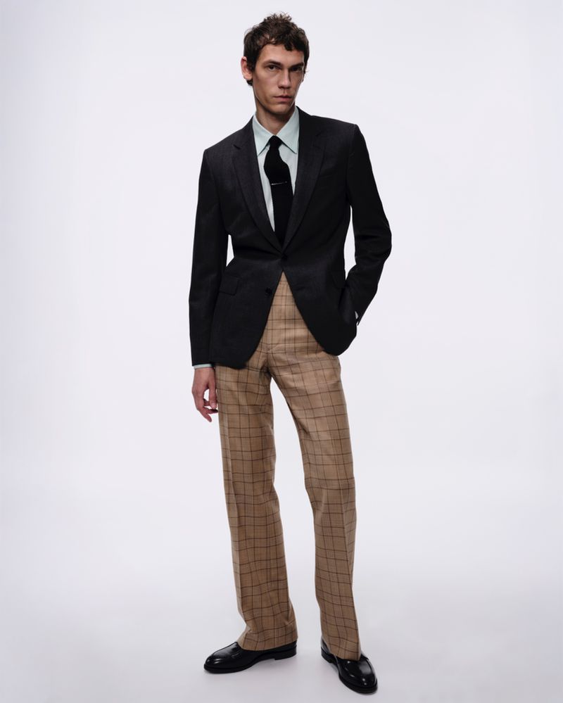 A man stands in a black blazer, light green shirt, and patterned dress pants against a clean, neutral backdrop in a full shot.