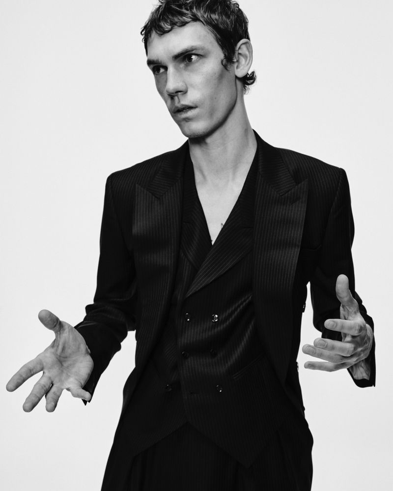 Monochrome portrait of a slender adult male in a tailored striped suit and vest, posing with outstretched hands against a bright white backdrop.