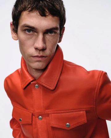 Close-up portrait of a young adult man in an orange leather jacket against a clean, white backdrop in a fashion setting.