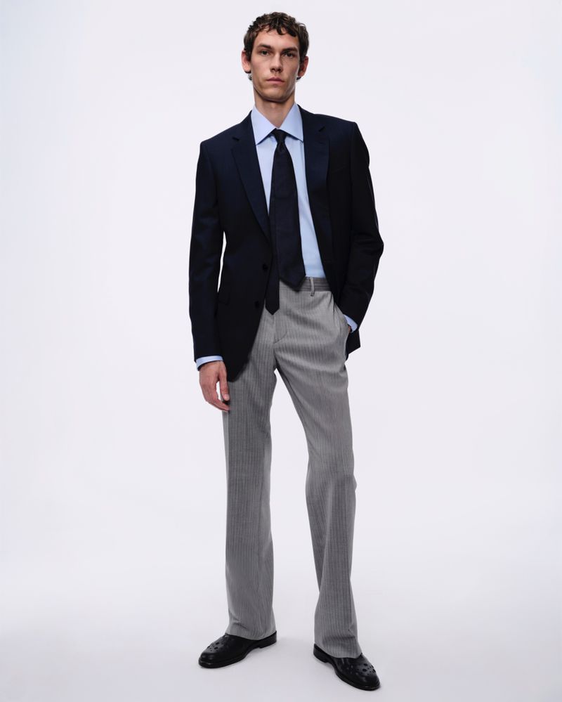 Full-length portrait of a male model wearing a dark blazer, light blue shirt, dark tie, and grey trousers against a seamless white backdrop.