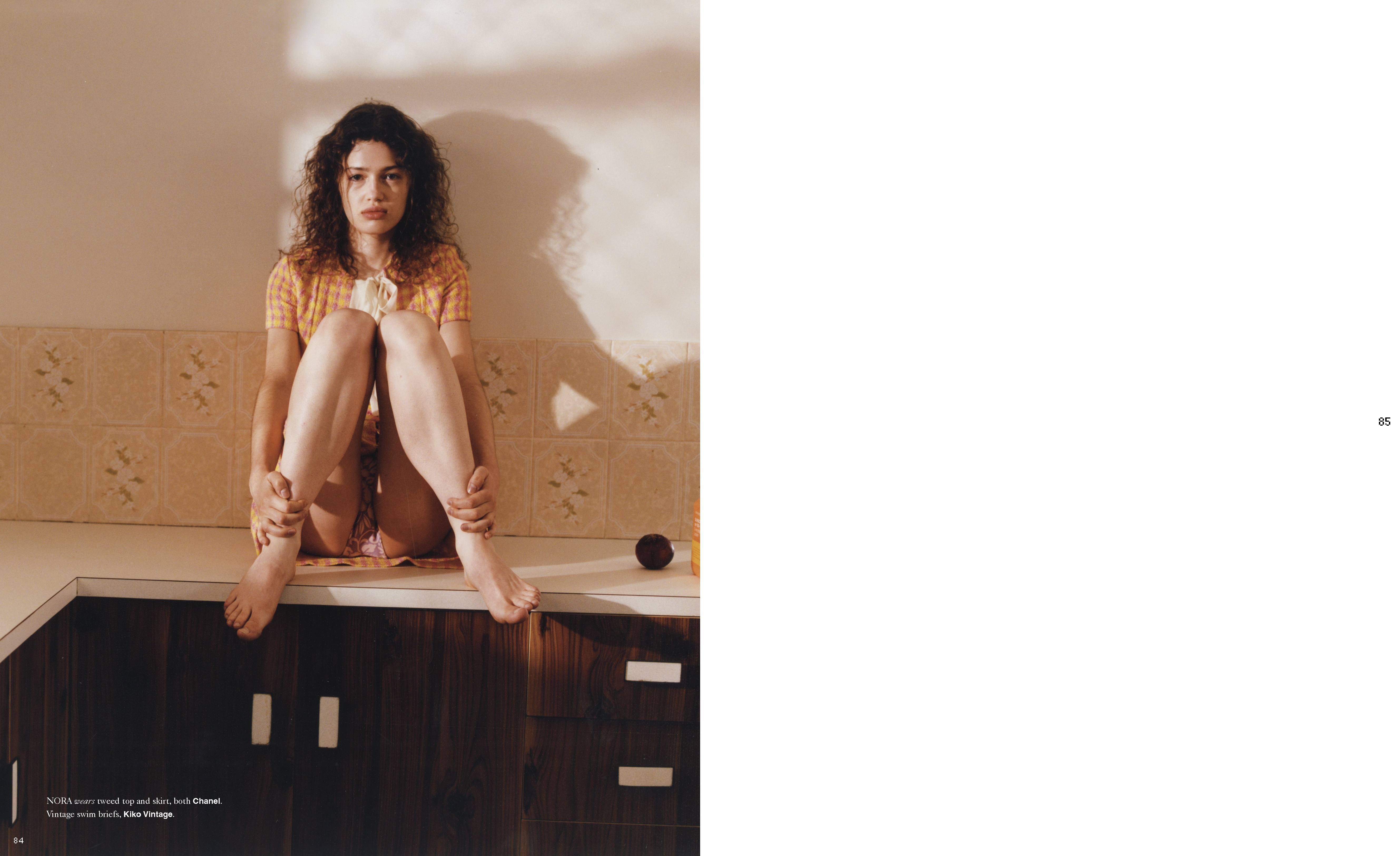 Young woman with curly hair sits on a countertop in a bright kitchen, hands hugging legs, wearing a patterned top. 