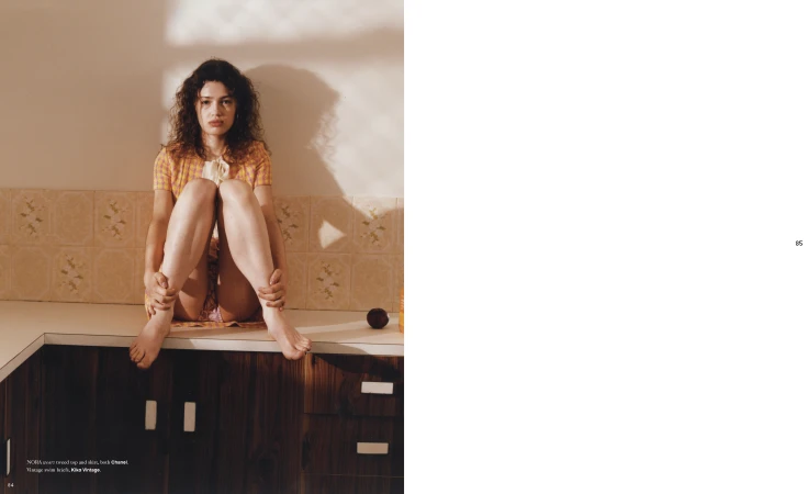 Young woman with curly hair sits on a countertop in a bright kitchen, hands hugging legs, wearing a patterned top. 