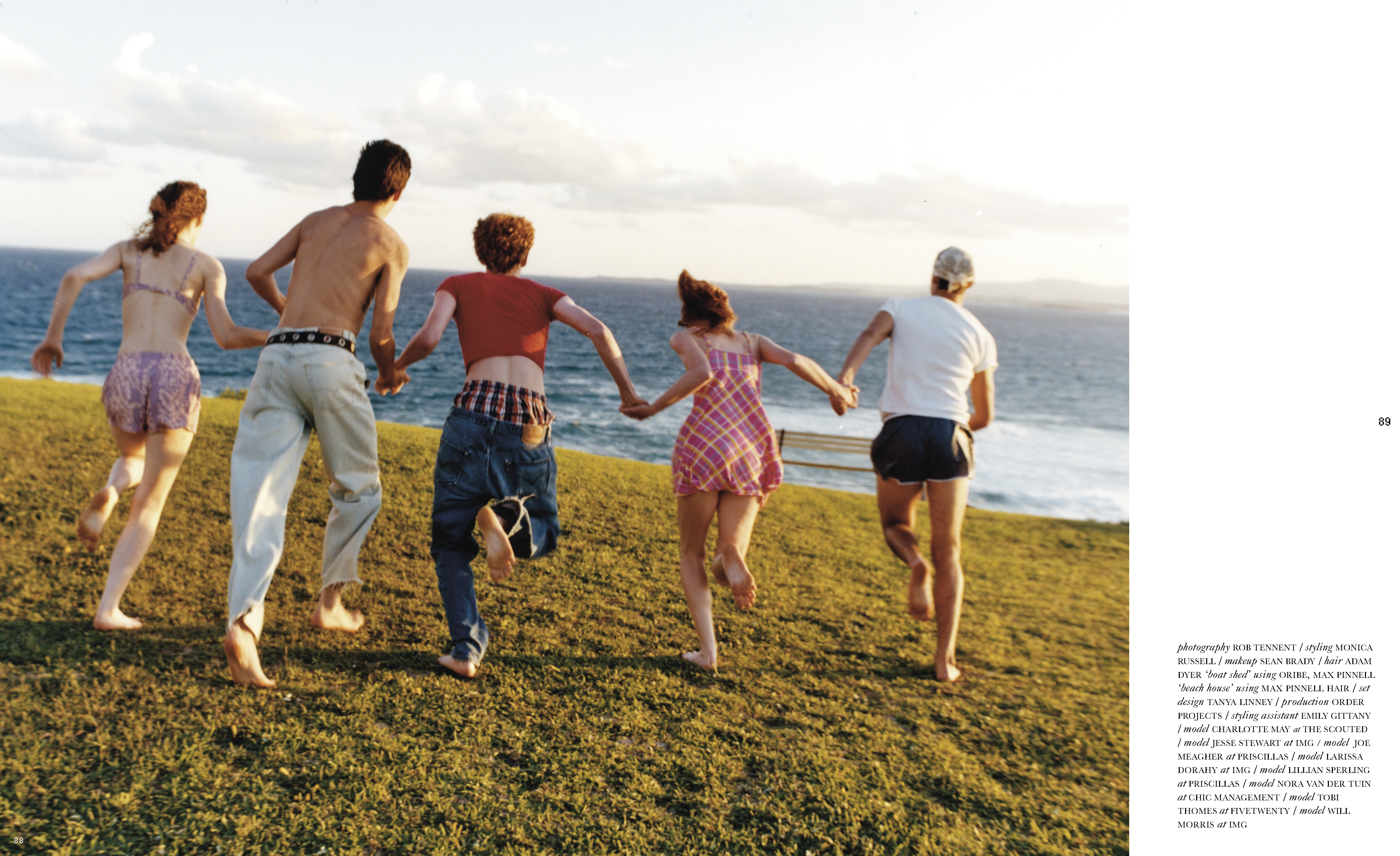 Five young adults run barefoot on a grassy hill towards the ocean under a bright sky, captured from behind, showcasing their casual summer attire.