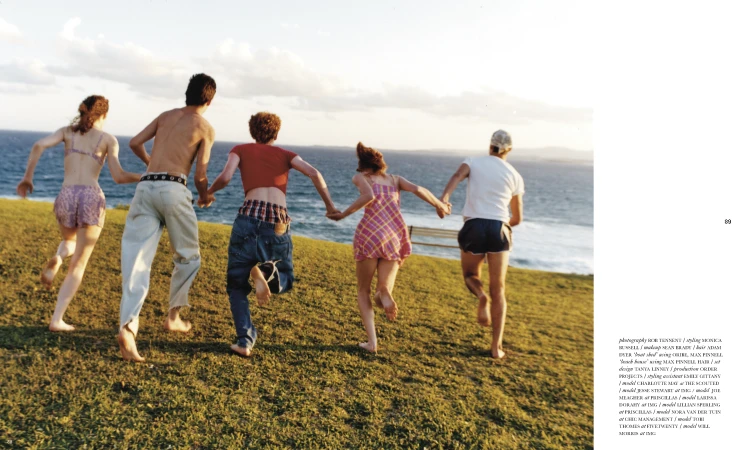 Five young adults run barefoot on a grassy hill towards the ocean under a bright sky, captured from behind, showcasing their casual summer attire.