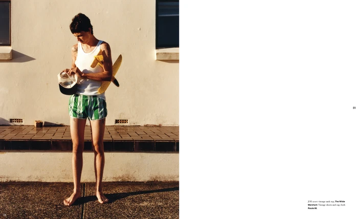 Young man in a white tank top and green striped shorts holding a cap and toy airplane against an outdoor wall.