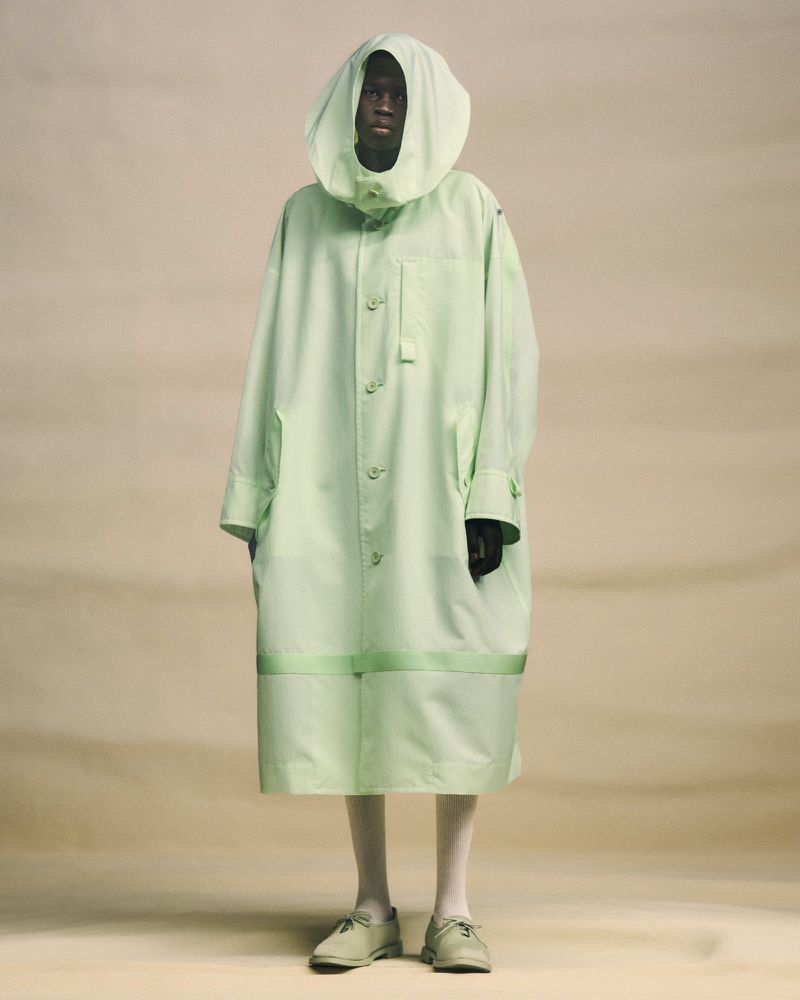 Dark-skinned adult in a hooded, light green oversized coat, white tights, and matching shoes poses against a neutral, soft background.