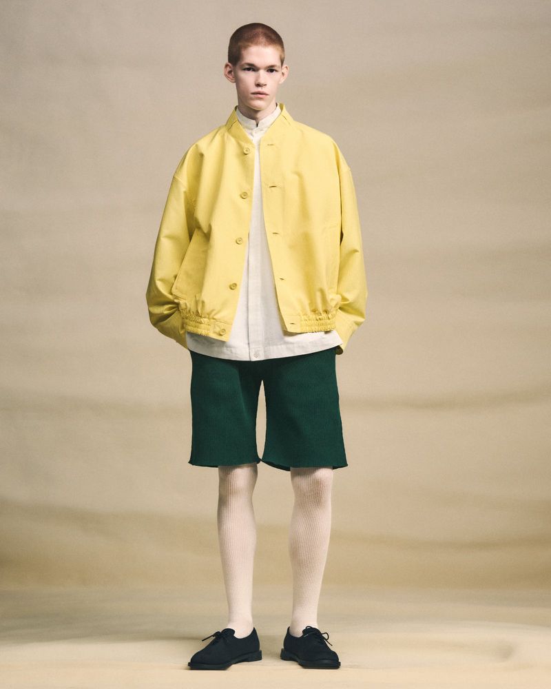A young adult fashion model poses in a studio wearing a yellow jacket, a white shirt, dark green shorts, and black shoes, looking forward.