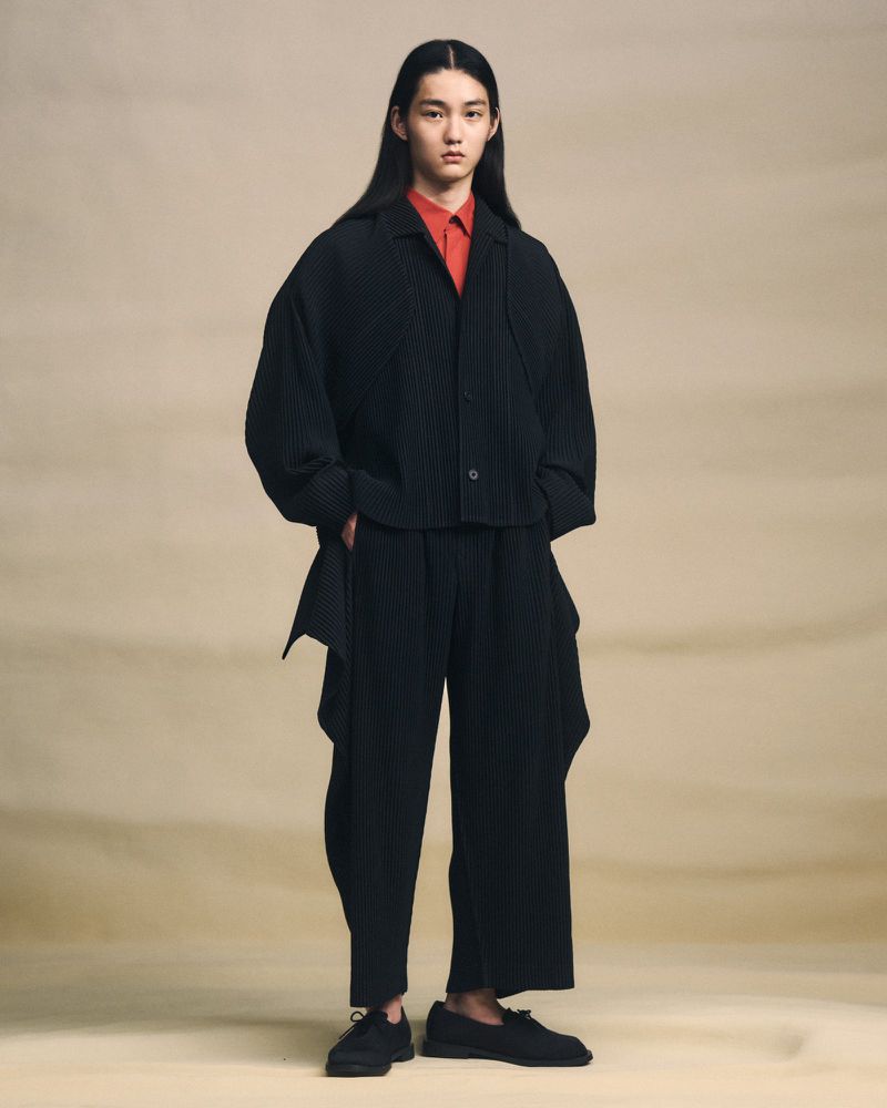 Young adult in dark, pleated jacket and pants, posing in a full shot against a neutral gradient backdrop, fashionably relaxed.