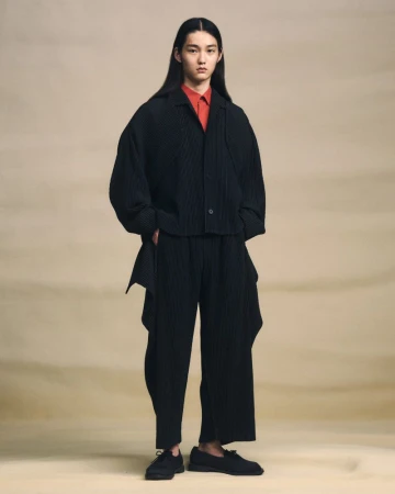 Young adult in dark, pleated jacket and pants, posing in a full shot against a neutral gradient backdrop, fashionably relaxed.