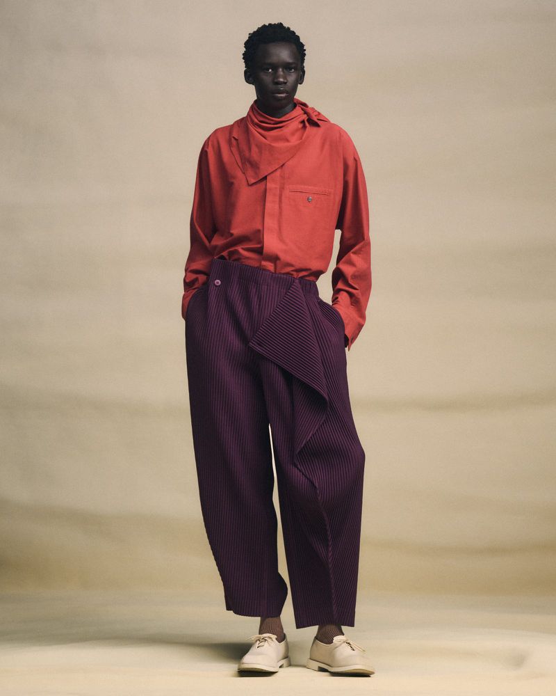 Fashion model wearing a red collared shirt with a neck tie and purple pleated pants in a full-length studio shot.