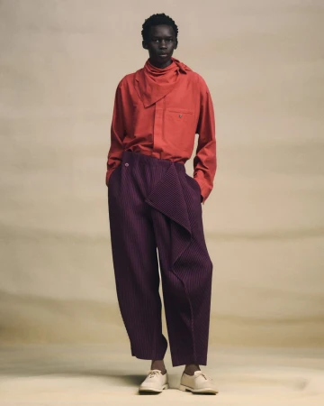 Fashion model wearing a red collared shirt with a neck tie and purple pleated pants in a full-length studio shot.
