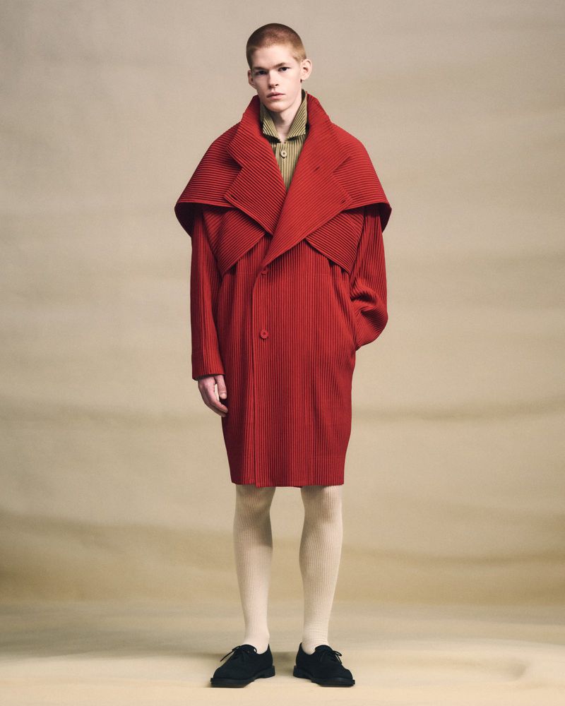 A young adult fashion model poses in a studio, wearing a red, ribbed coat, cream-colored tights, and black shoes against a neutral backdrop.