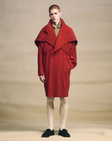 Model wearing a pleated red overcoat standing against a neutral textured studio backdrop.