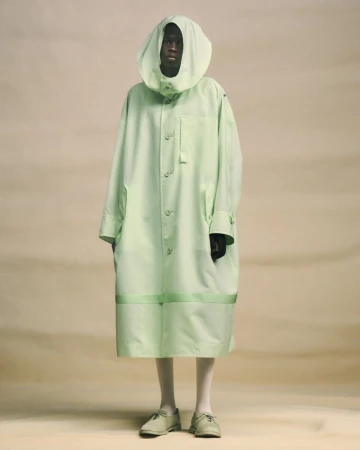 Model wearing a light green hooded coat and matching shoes against a neutral studio backdrop for Issey Miyake.
