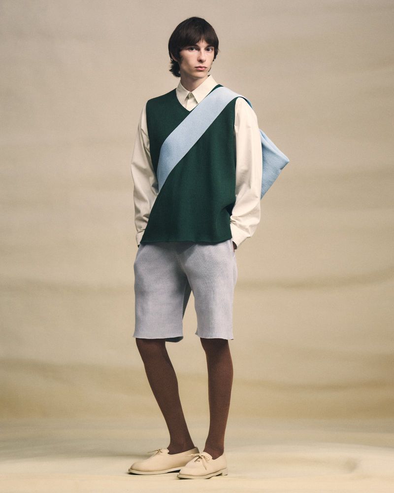 Model standing against a neutral textured background wearing a green sweater vest over a white shirt and light shorts