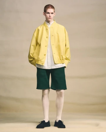 Model wearing a yellow jacket and green shorts against a neutral studio backdrop for Issey Miyake.