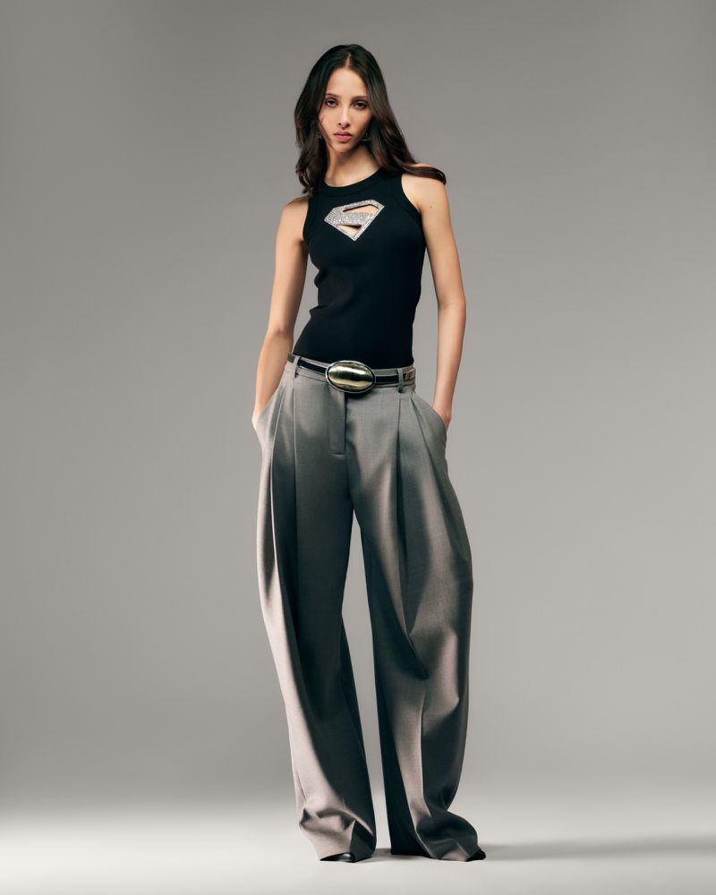 Fashion shot of a young woman with long brown hair, wearing a black top with a Superman logo and wide grey pants, in a studio setting.