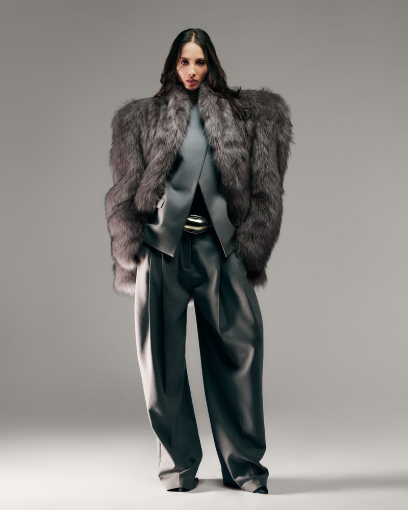 A female fashion model poses in a grey fur coat, grey top, wide-legged grey trousers, and a silver belt against a neutral backdrop.