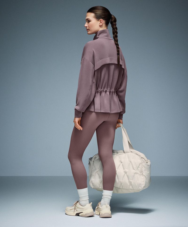 Woman in mauve athletic wear with a white quilted gym bag, seen from the back in a studio shot with a blue-grey gradient background.