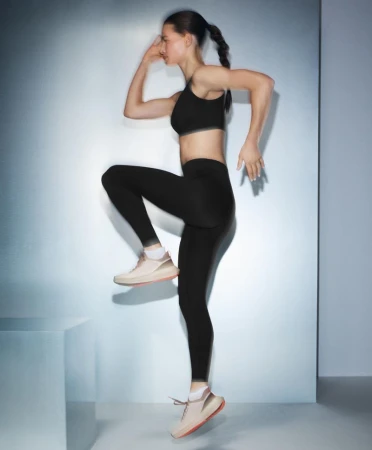 A woman in black athletic wear and sneakers is exercising indoors in front of a silver wall; motion blur is visible.