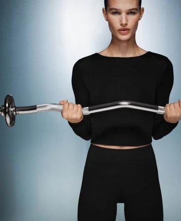 Athletic woman in fitness apparel lifts a silver EZ curl bar in a photo studio with a blue and white gradient background.