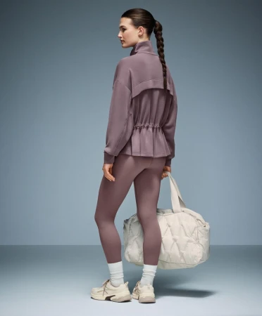 Model in mauve activewear set with white sneakers holding a quilted duffel bag against a gradient grey studio background.
