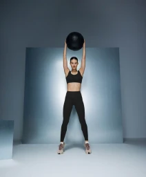 Model in black activewear holding a medicine ball above her head in a studio setting for Oysho training project