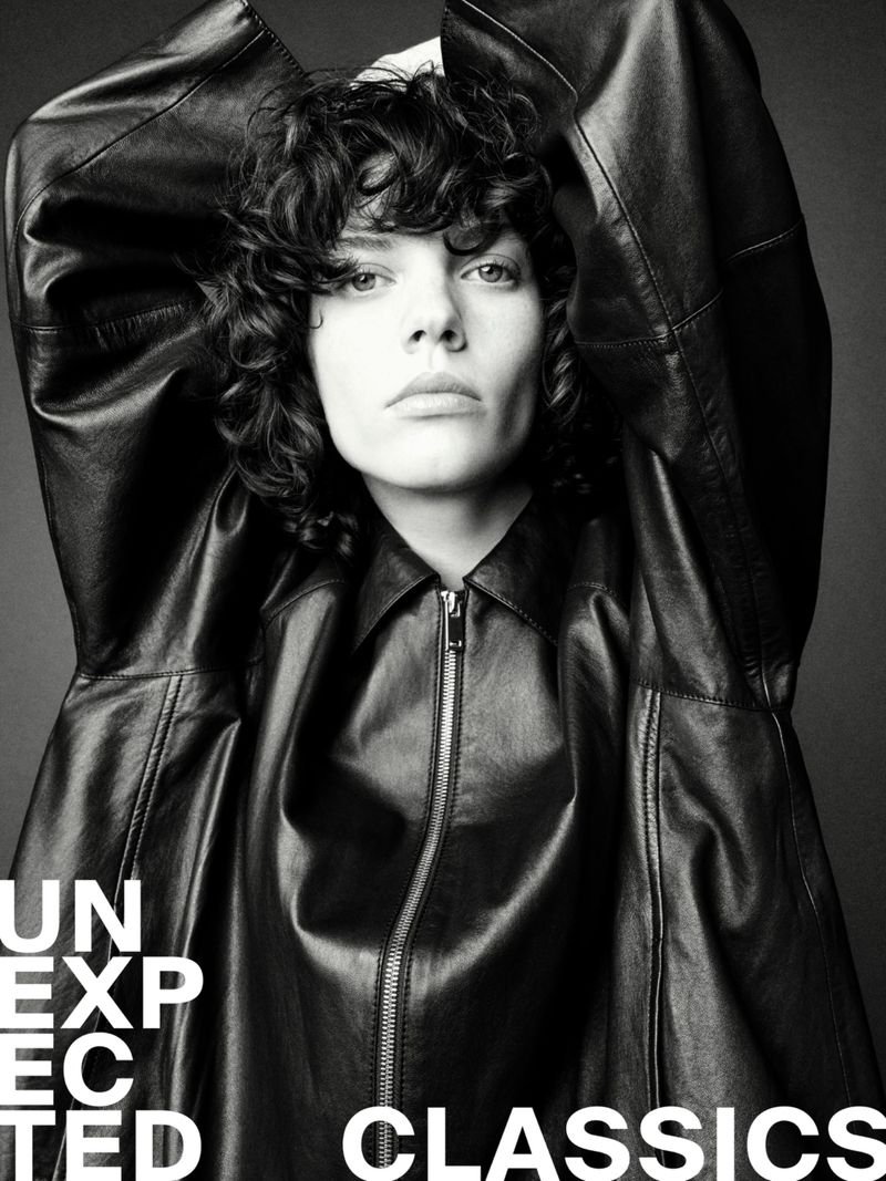 Close-up black and white portrait of a person with curly hair in a leather jacket looking at the camera with a neutral expression.