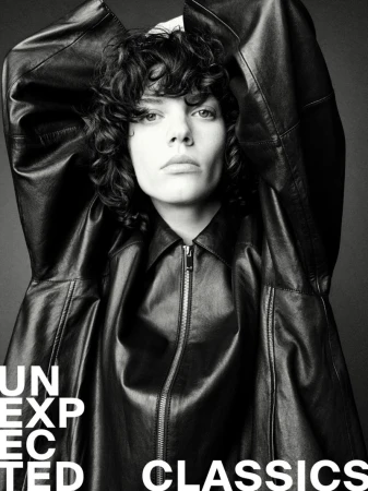 Close-up black and white portrait of a person with curly hair in a leather jacket looking at the camera with a neutral expression.