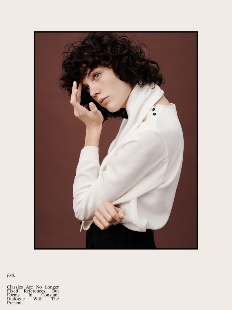 Editorial fashion shot of woman with curly hair wearing white blouse against brown backdrop, shot in a studio with natural lighting.