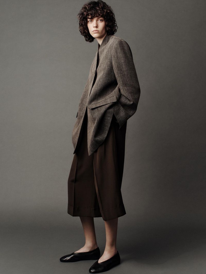 Fashion shot of a woman wearing a brown blazer, culottes, and black flats, posing in front of a gray backdrop in a studio.