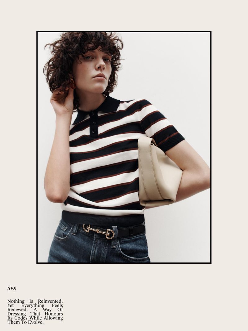 A woman with curly hair poses in a striped polo shirt and jeans, holding a beige purse in an indoor setting with a light background.
