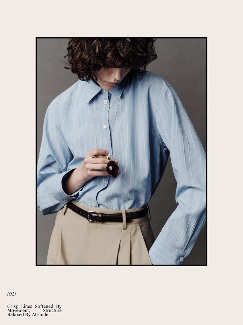 A person poses indoors wearing a blue striped button-down shirt, tan pleated pants, and a black belt while holding a small perfume bottle.