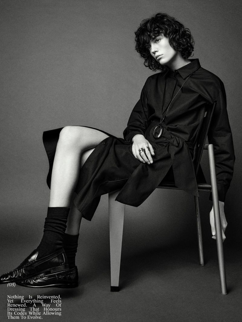 Monochrome fashion shot of a curly-haired model in a black outfit seated on a chair in a studio with soft lighting, showcasing an elegant, minimalist aesthetic.