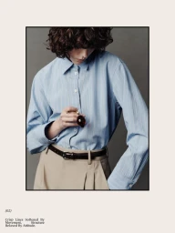 Model wearing a light blue striped shirt and beige trousers with a thin dark belt, holding a pendant necklace.