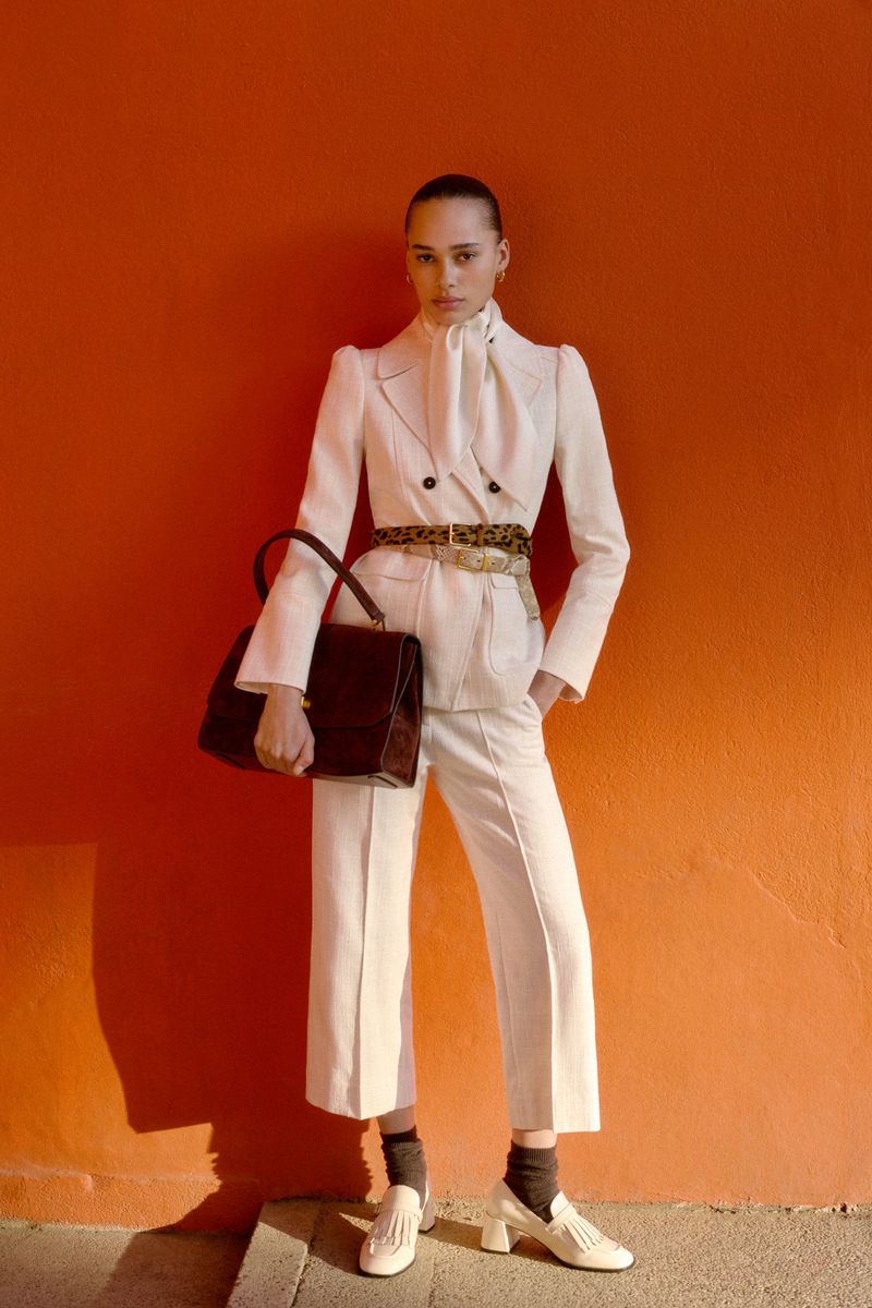 Woman poses in white pantsuit with brown bag against orange wall in eye-level shot, showcasing fashion editorial style with natural light.