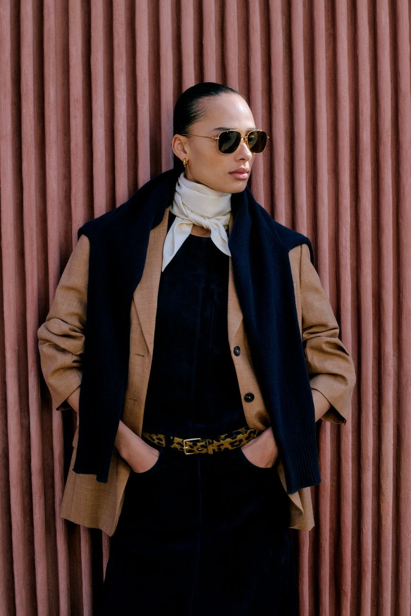 Stylish woman in sunglasses, tan blazer, and scarf, posing against a textured pink wall, showcasing fashion and portrait photography.