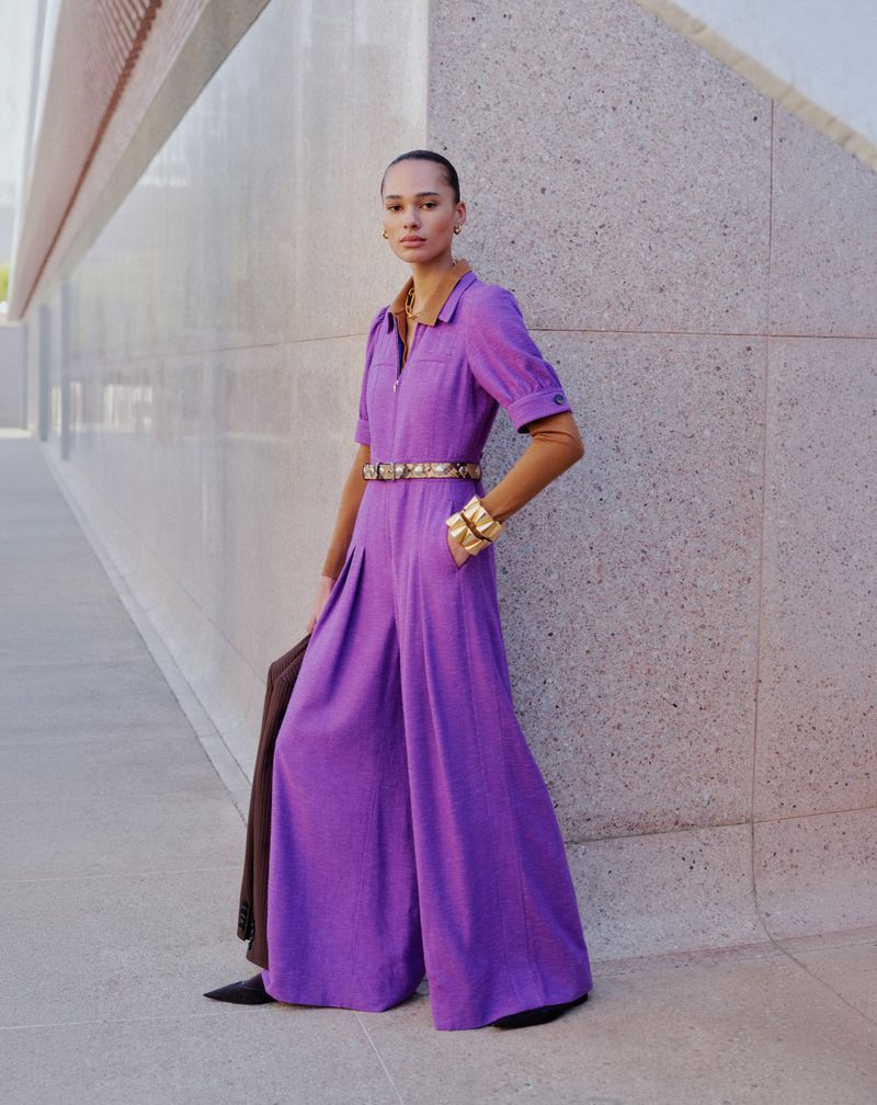 Elegant woman in a purple jumpsuit with gold accessories stands in front of a modern building for a fashion editorial shoot.